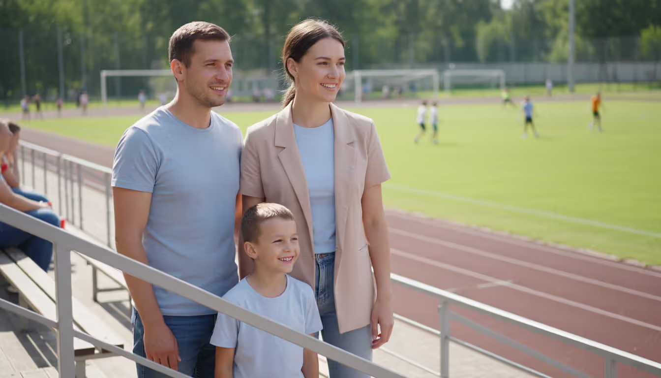 A man and a woman standing together on school bleachers watching their child play sports, both looking supportive and friendly in a sunny outdoor setting