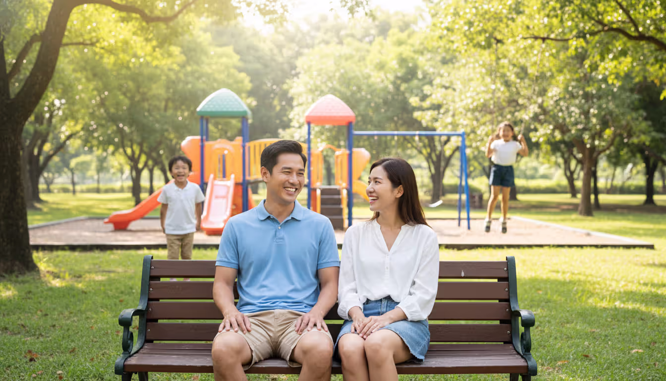 Two parents sitting on a park bench smiling while watching their two children playing on a playground on a sunny day